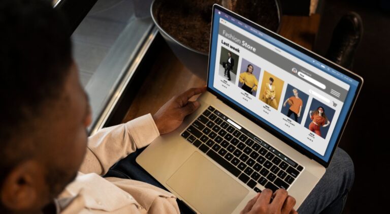 Homem negro, com camisa clara, sentado e digitando em um laptop. A tela do computador exibe a página de uma loja de roupas online chamada 'Fashion Store', com várias fotos de modelos exibindo peças de vestuário.