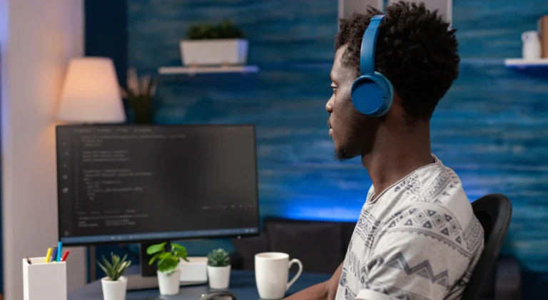 Um homem negro jovem, visto de perfil, sentado em frente a um computador em um escritório com parede azul ao fundo. Ele usa fones de ouvido azuis grandes e uma camiseta cinza com estampas geométricas, e olha atentamente para o monitor que exibe linhas de código em fundo escuro.