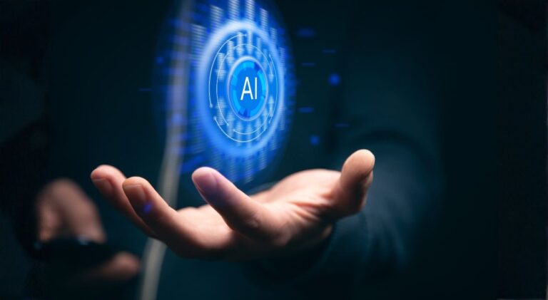 Mão estendida segurando uma projeção holográfica azul brilhante de um círculo tecnológico com as letras "AI" (Inteligência Artificial) no centro. A imagem tem um fundo escuro e foca na interface digital futurista, simbolizando a IA nas mãos humanas.