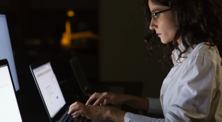 Mulher branca, com camisa branca, óculos preto e cabelos escuros olhando para tela do notebook e digitando no teclado.