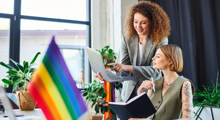 Duas mulheres sorridentes trabalhando juntas em um notebook em um escritório. Em primeiro plano, desfocada, há uma bandeira do arco-íris LGBTQIA+.