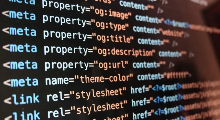 Close em uma tela de computador exibindo linhas de código de programação HTML em um editor de texto com fundo escuro. As tags e estão em destaque com cores azul-claro, branco e laranja, mostrando propriedades de Open Graph como "og:image", "og:type", "og:title", "og:description", além de "theme-color" e caminhos para folhas de estilo ("stylesheet"). A imagem está levemente inclinada e possui um efeito de profundidade, deixando o lado direito do código um pouco desfocado.