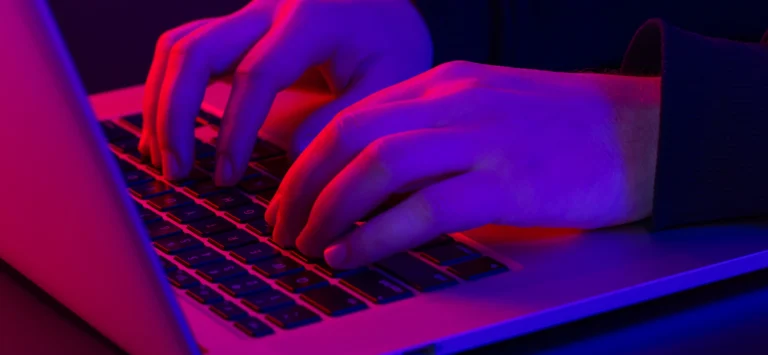 Close em mãos digitando no teclado de um notebook, iluminadas por luzes neon nas cores azul, roxo e vermelho.