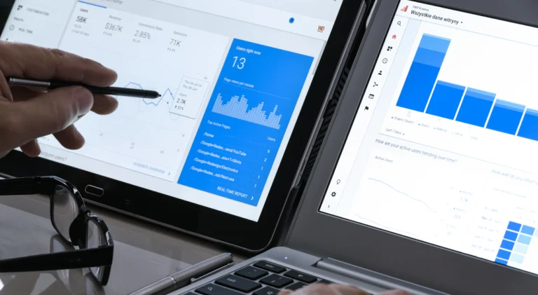 Mão com caneta aponta para gráficos e dados do Google Analytics em um tablet ao lado de um notebook.