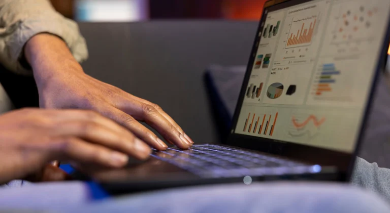 Mãos digitando em notebook com dashboard de gráficos e métricas exibido na tela.