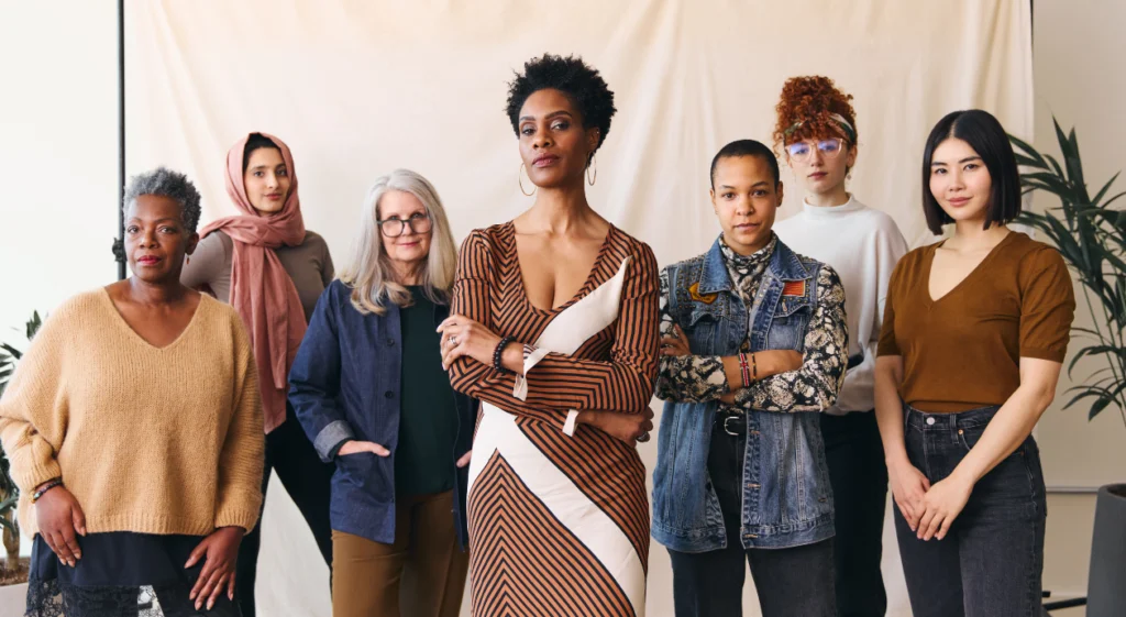 Foto em plano médio de sete mulheres com diversas etnias, idades e estilos, de pé lado a lado olhando confiantemente para a câmera contra um fundo neutro.