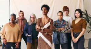 Foto em plano médio de sete mulheres com diversas etnias, idades e estilos, de pé lado a lado olhando confiantemente para a câmera contra um fundo neutro.