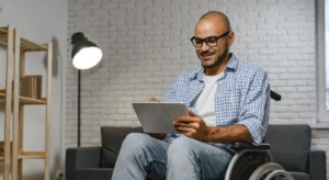 Homem cadeirante sorrindo enquanto usa um tablet, em ambiente iluminado com sofá e estante ao fundo.