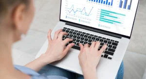 Pessoa digitando em notebook com dashboard de gráficos de linha e barras em tons de azul na tela.