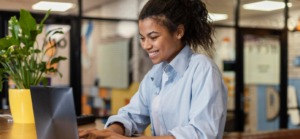 Mulher jovem sorridente de camisa azul trabalha no notebook em um ambiente de escritório moderno e iluminado.