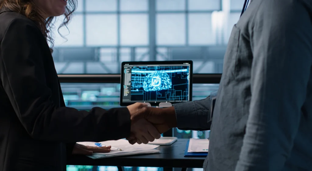 Duas pessoas apertando as mãos em ambiente corporativo, com um monitor ao fundo exibindo a ilustração de um cérebro digital, representando uma parceria em torno de inteligência artificial.