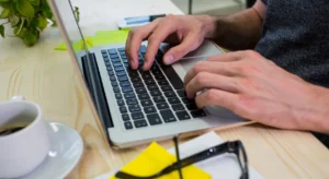Mãos digitando em notebook sobre mesa com post-its amarelos, óculos e xícara de café ao redor.